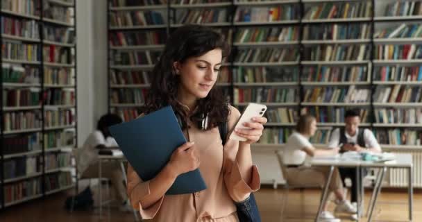 Attraktive Studentin steht mit Handy in Bibliothek — Stockvideo