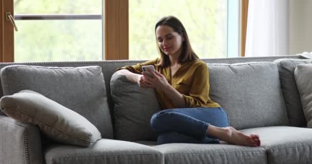 Ung kvinna sitta på soffan håll telefonen njuta av att dejta online — Stockvideo