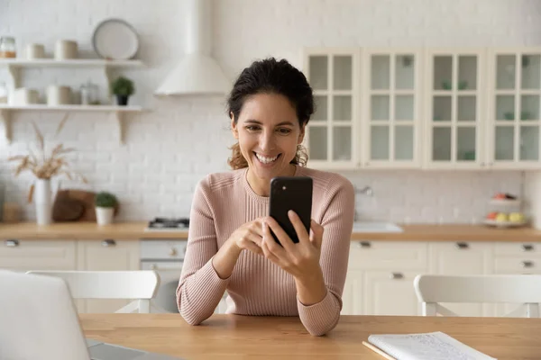 Mujer emocionada sosteniendo teléfono inteligente, charlando en línea en la cocina moderna — Foto de Stock
