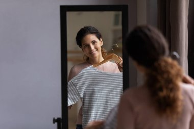 Gülümseyen bir kadının aynadaki yansıması elbise deniyor, askı tutuyor.