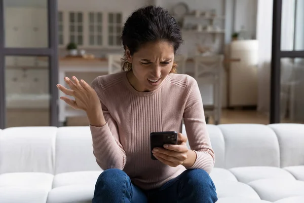 Wanita marah bingung melihat layar smartphone, duduk di sofa — Stok Foto