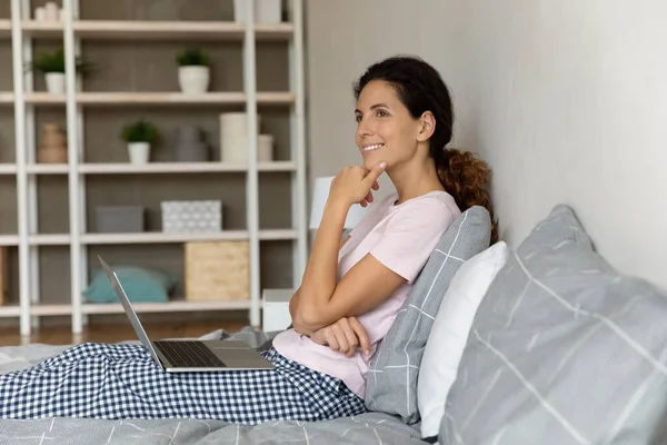 Mujer sonriente y soñadora distraída de la computadora portátil, sentada en la cama — Foto de Stock