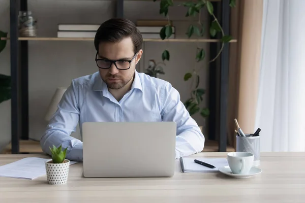 Молодий серйозний бізнесмен сидить за столом, працює на ноутбуці — стокове фото