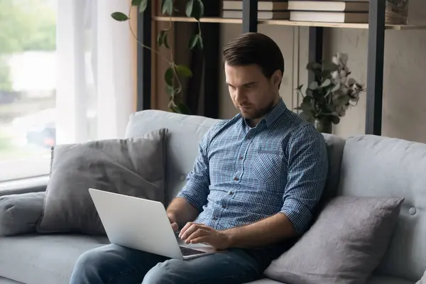 Серйозний зосереджений чоловік сидить на дивані вдома з ноутбуком — стокове фото