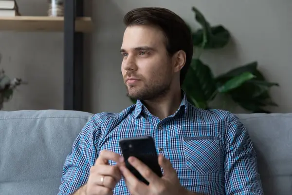 El hombre se sienta en el sofá sostiene teléfono inteligente mirando a la distancia — Foto de Stock