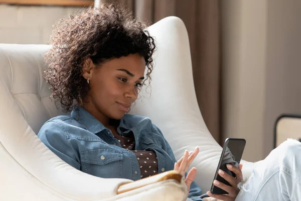 Atractiva mujer africana calma relajante en acogedor sillón con teléfono inteligente — Foto de Stock