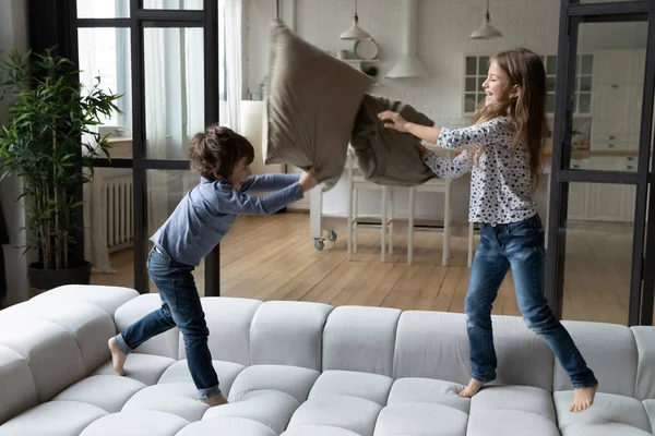 Adik perempuan dan saudara laki-laki yang gembira bertengkar bantal, melompat-lompat di sofa — Stok Foto