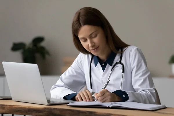 Geconcentreerde jonge vrouw medisch medewerker neemt notities op de werkplek — Stockfoto