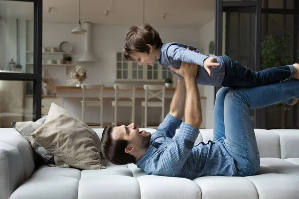 Glücklicher junger Vater liegt auf Couch und hebt aufgeregten Sohn — Stockfoto