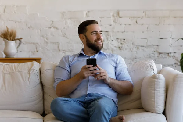 Glad drömsk ung man som använder smartphone hemma. — Stockfoto