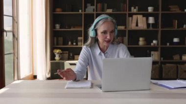 Kulaklıklı yaşlı bayan öğretmen laptopla öğrenciye danışıyor.