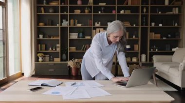 Yaşlı iş kadını, kağıt belgelerindeki istatistikleri kullanarak dizüstü bilgisayarda çalışıyor.