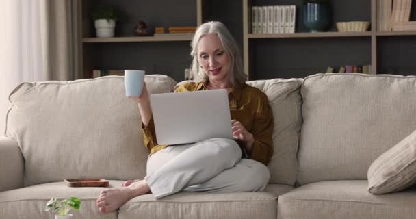 Babička relaxovat doma s notebookem drží šálek čaje — Stock video