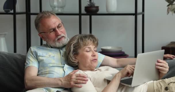 Senior spouses rest on sofa browse media content on laptop — Video Stock