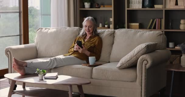 Beautiful senior adult female sit on sofa use mobile phone — 图库视频影像