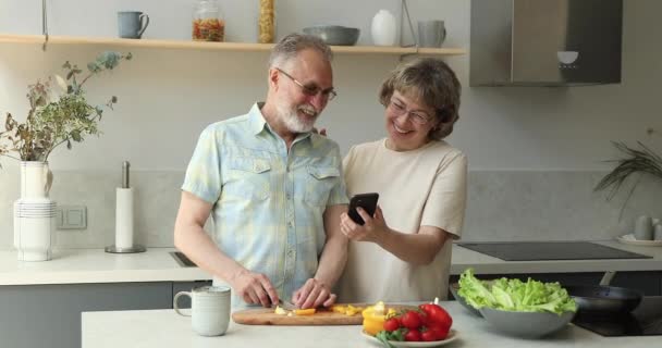 Happy ældre mand lave mad middag snak med kone holder telefonen – Stock-video