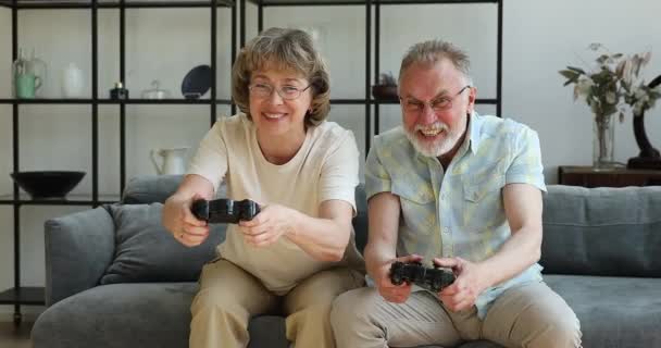 Ενθουσιασμένο ηλικιωμένο ζευγάρι κάθεται στον καναπέ και απολαμβάνει να παίζει κονσόλα βιντεοπαιχνίδι — Αρχείο Βίντεο
