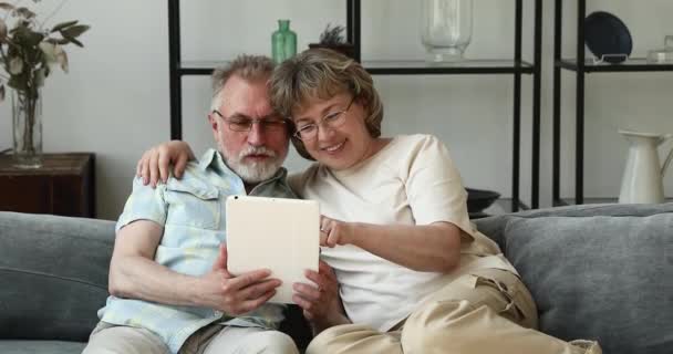 Χαμογελαστό ηλικιωμένο ζευγάρι ξεκουράζεται στον καναπέ χρησιμοποιώντας touchpad διασκεδάζοντας — Αρχείο Βίντεο