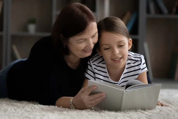 Щаслива школярка насолоджується читанням смішної книги разом з бабусею у віці — стокове фото
