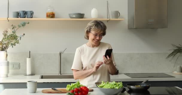 Oudere vrouw afgeleid van koken lees culinair recept met behulp van telefoon — Stockvideo