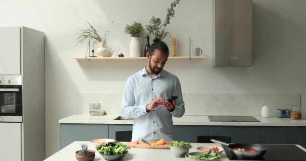 Afrikalı adam cep telefonuyla yemek tarifi veriyor, mutfakta yemek yapıyor. — Stok video