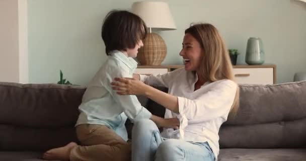 Joyful young female babysitter tickling on sofa with little boy — стоковое видео
