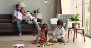 Genç aileler kanepede dinlenir, çocukları yerde oynarken izlerler.