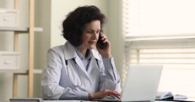 Yaşlı doktor dizüstü bilgisayarla ilgileniyor. Hastayla cep telefonundan konuşuyor.