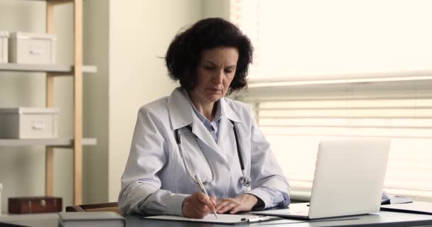 Older female general practitioner writing, using laptop, working at workplace — ストック動画