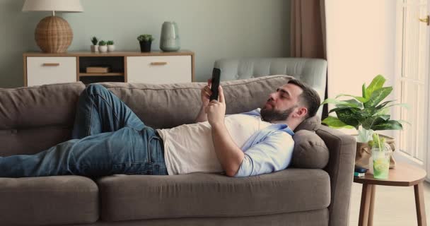 Relaxed millennial guy lie on couch watch video on phone — Stockvideo