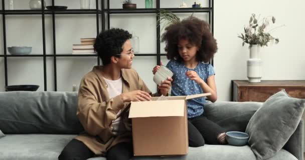 African girl her mother unpack parcel box with diverse kitchenware — Stockvideo