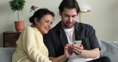 Yaşlı kadın, yetişkin oğlu kanepeye oturur ve akıllı telefon kullanır.