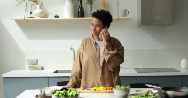 African woman talks on phone while cooking salad in kitchen — Vídeo de Stock