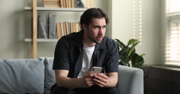 Millennial guy sit on sofa holds cellphone chatting with friend — Stockvideo