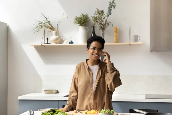 Chica afroamericana feliz hablando por teléfono celular mientras cocina la cena —  Fotos de Stock