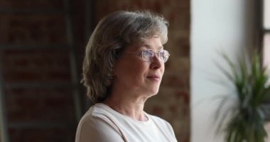 Gri saçlı, gözlüklü yaşlı kadın içeride dikilip yan yana bakıyor.