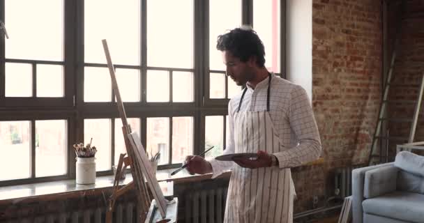 若いヒスパニック系男性画家は、スタジオでキャンバス上の絵画を作成します — ストック動画