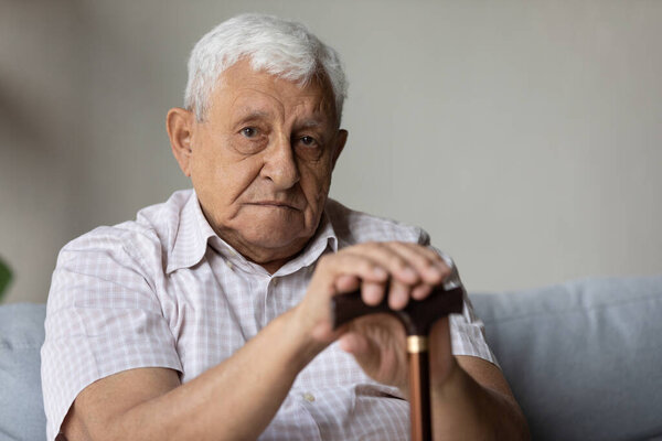Portrait of upset old pensioner with wooden cane in hands.