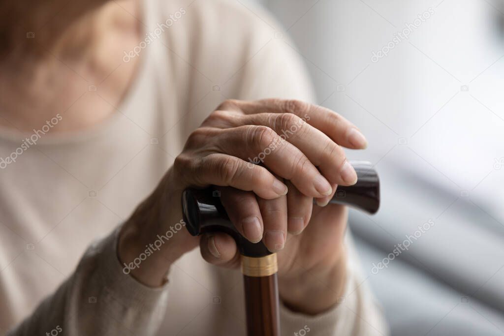 Close up focus on folded wrinkled female hands on wooden cane.