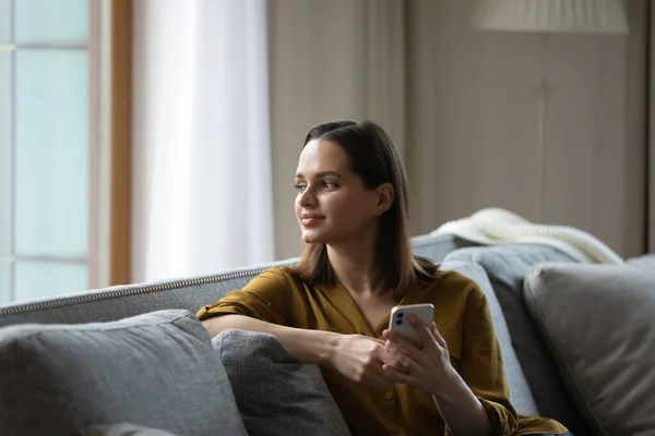 Happy dreamy millennial girl holding smartphone, resting on comfortable sofa — Stock Photo, Image
