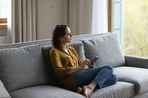Thoughtful serious millennial girl using smartphone, sitting on comfortable sofa — Stock Photo, Image