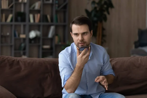 Hombre serio grabando mensaje de audio en el teléfono inteligente, sentado en el sofá — Foto de Stock