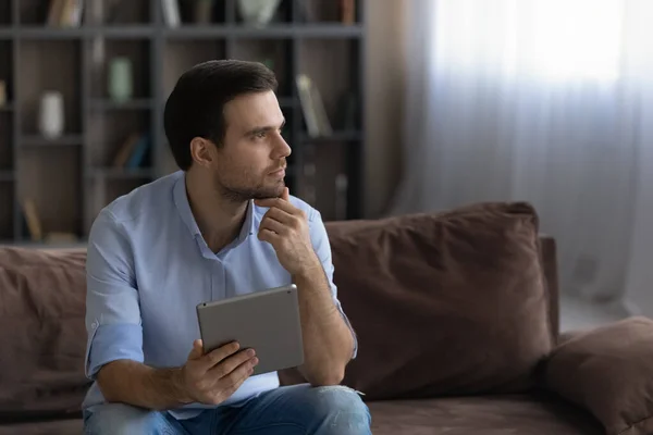 Thoughtful man holding tablet, touching chin, looking in distance — ストック写真