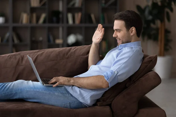 Profile view smiling man waving hand, using laptop on couch — 图库照片