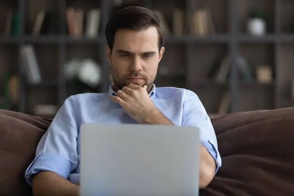Thoughtful man looking at laptop screen, touching chin, working online — ストック写真