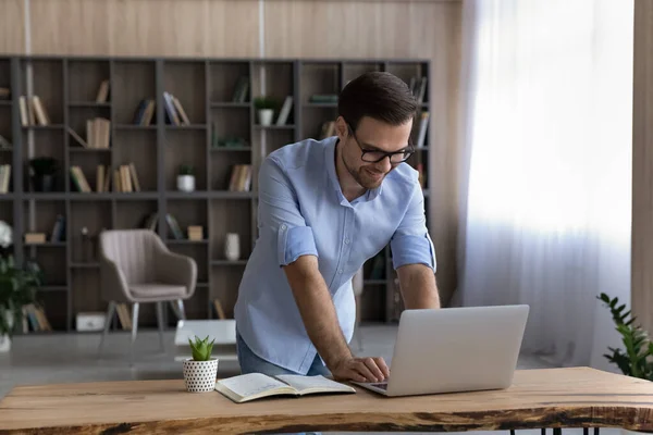 Smiling businessman in glasses using laptop, standing at desk — Stock Photo, Image