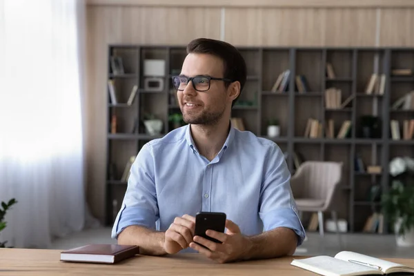 Sorridente uomo d'affari sognante indossa occhiali guardando in lontananza, tenendo smartphone — Foto Stock