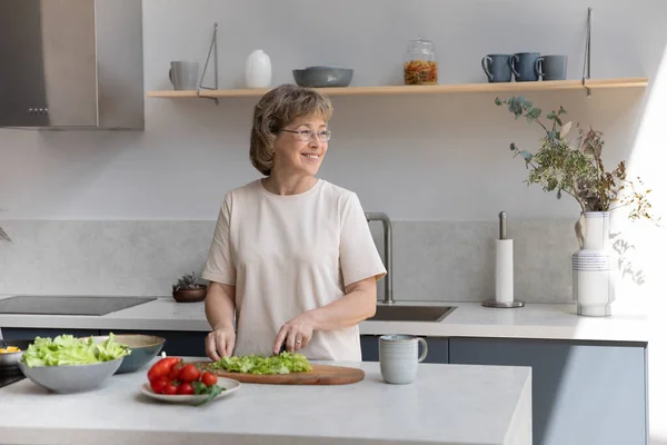 Sonriente mujer de mediana edad soñadora cocinando en la cocina. —  Fotos de Stock