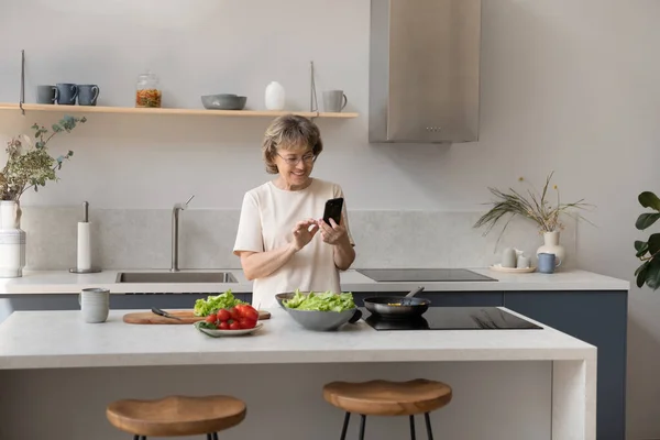 Feliz anciana de mediana edad utilizando el teléfono celular preparando comida. — Foto de Stock
