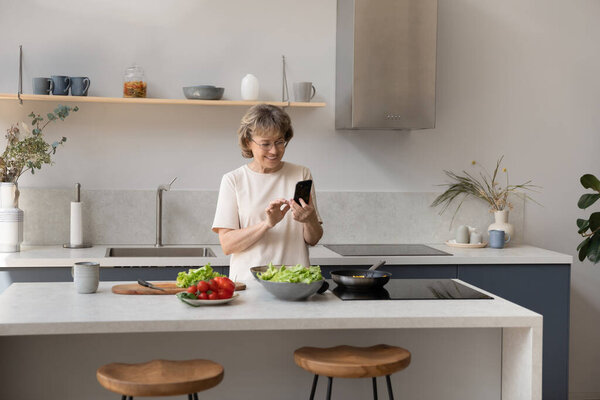 Happy middle aged old woman using cellphone preparing food.